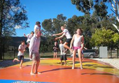 A group of children and adults enjoying a game on an inflatable jumping pillow, set against a backdrop of trees and clear blue skies.