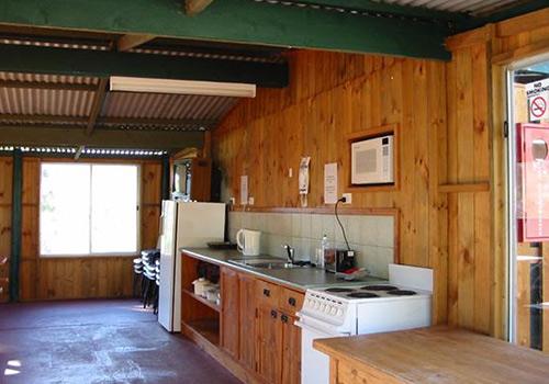 A cozy kitchen area with wooden walls, a refrigerator, microwave, stove, and sink, featuring ample storage space and natural light streaming through large windows.