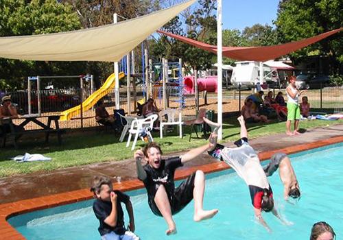 A lively scene at a swimming pool where children are jumping into the water, with colorful playground equipment and shaded seating areas in the background.
