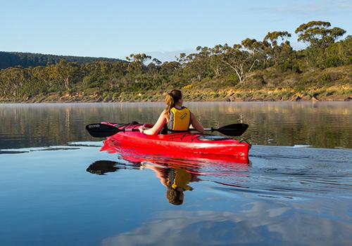 A kayaker paddling a red kayak on calm waters, surrounded by lush green hills and reflecting the serene beauty of the landscape.