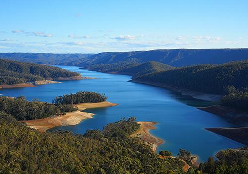 A breathtaking view of a serene lake surrounded by lush green hills and dense forests, with calm waters reflecting the clear blue sky.