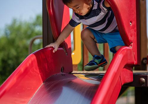 A young child climbing up a red slide at a playground, wearing a striped shirt and blue shorts.