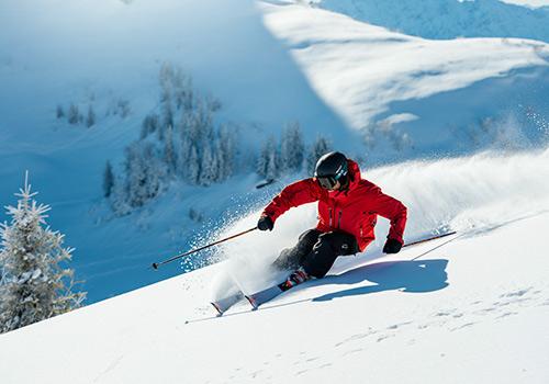 A skier in a red jacket glides down a snowy slope, leaving a trail of snow spray behind, with a backdrop of snow-covered trees and bright sunlight.