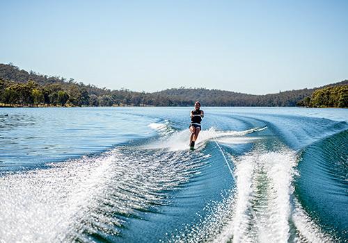 A person water skiing on a calm lake, leaving a trail of white spray as they glide across the water.